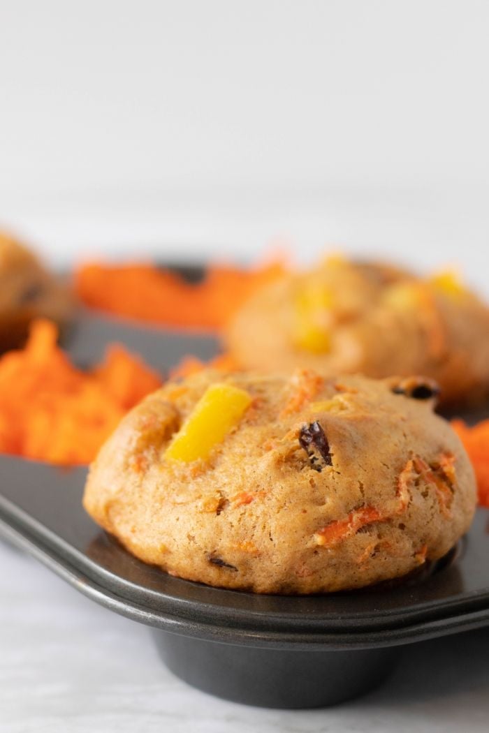 a carrot pineapple muffin sits in a muffin tray, lots of shredded carrot can be seen in the background